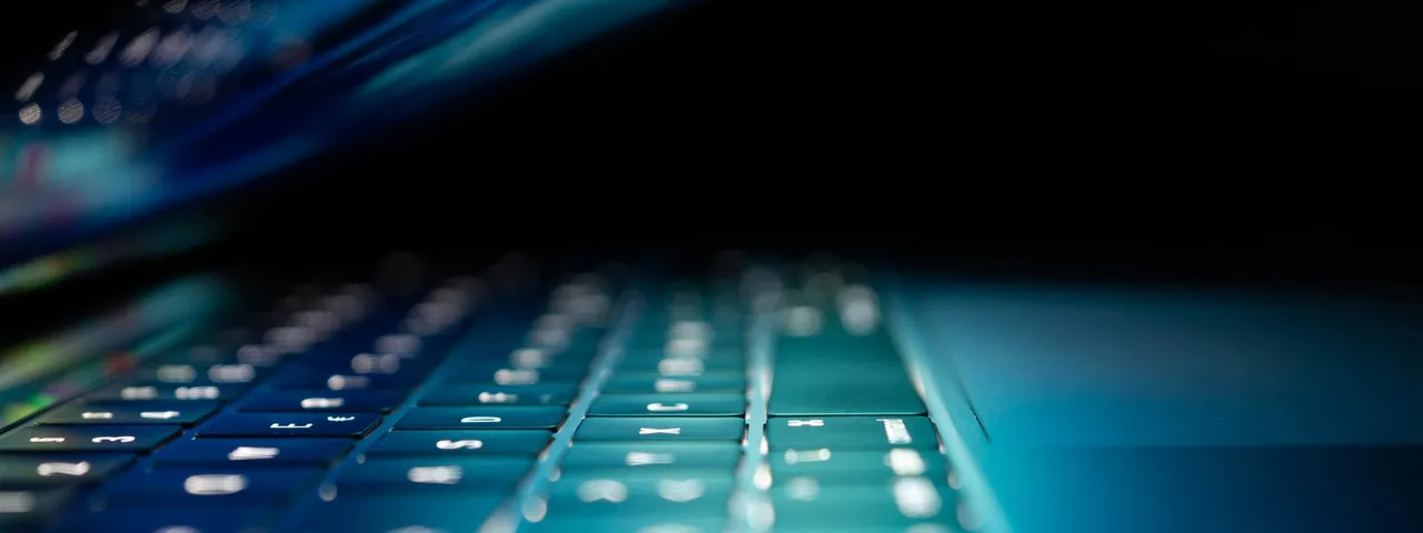 A backlit laptop keyboard glowing blue in a dark room, illustrating bulk distribution list migration to Exchange Online with PowerShell.